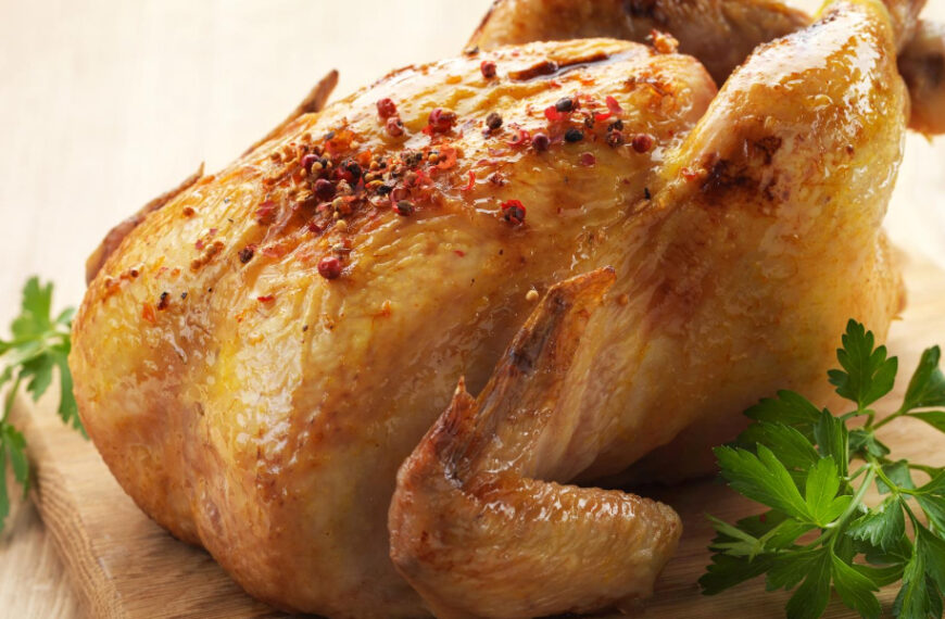 A roasted chicken garnished with red pepper flakes is placed on a wooden board, accompanied by sprigs of parsley.